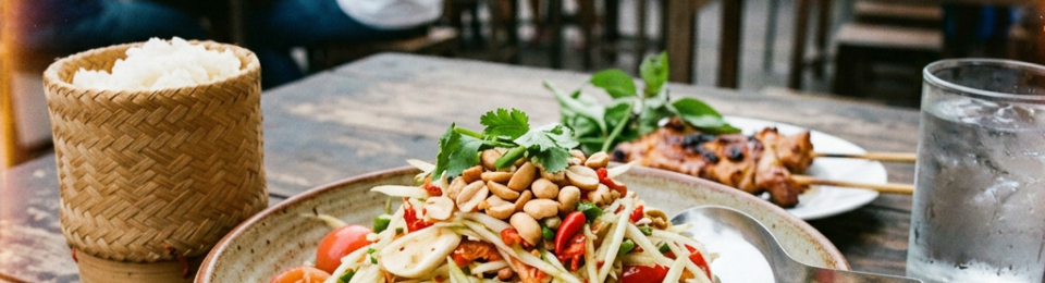 Thai papaya salad with peanuts, lime, and shrimp with sticky rice and grilled skewers on wooden table
